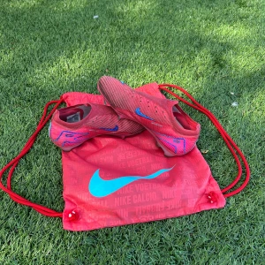 Röda Nike fotbollsskor med blå detaljer - Säljer ett par röda Nike fotbollsskor med blå swoosh och grafiska detaljer. Skorna har en smal passform och är designade för att ge bra grepp på planen. Kommer med en matchande röd Nike-påse med blå logga. Perfekta för dig som vill sticka ut på fotbollsplanen.