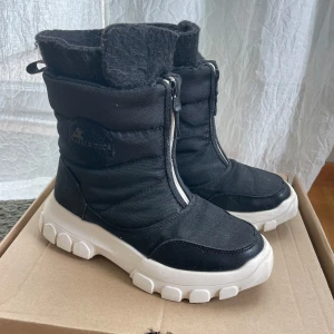 Svarta vinterboots från Rubber Duck - Svarta vinterboots från Rubber Duck med vit, rejäl sula och dragkedja framtill. Skorna är vadderade och har en sportig look med rund tå och grovt mönstrad yttersula. Perfekta för kalla dagar och enkel att ta på tack vare dragkedjan.