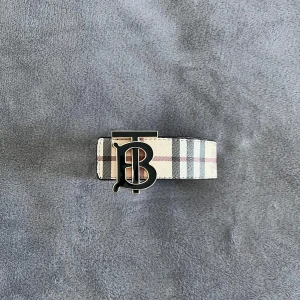 Burberry bälte  - Snyggt tvåsidig Burberry-bälte med ikoniskt rutmönster i beige, svart, vitt och rött. Bältet har en stor metallspänne med TB-logga och är tillverkat i skinn. Perfekt accessoar för att lyfta din outfit med en lyxig touch.