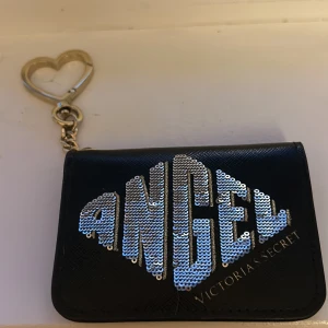 Svart plånbok Victoria's Secret Angel - Svart plånbok från Victoria's Secret med texten 'ANGEL' i silvriga paljetter på framsidan. Plånboken har ett hjärtformat nyckelringsfäste i metall, flera kortfack (4 stycken)och ett fönster för ID-kort på baksidan. Snygg och praktisk accessoar med exklusiv känsla. Går inte att köpa på victoria secret nu,plånboken är då vintage men väldigt bra skick (inga synliga skador eller något sånt) Köpt för 250/300 säljer då för 200kr+frakt priset kan diskuteras vid snabbt och smidigt köp!
