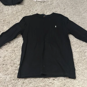 Svart långärmad tröja Polo Ralph Lauren - Svart långärmad tröja från Polo Ralph Lauren i storlek XL junior. Tröjan har en klassisk rund hals och är tillverkad i mjuk bomull. På bröstet finns den ikoniska broderade loggan i vitt. Perfekt för dig som gillar stilrena och tidlösa plagg.