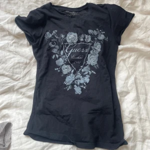 Svart blommig t-shirt från Guess - Svart t-shirt från Guess med korta ärmar och tryck framtill med blommor och Guess-logga. Vissa blommor har paljettdetaljer som ger extra bling. Snygg passform och mjukt bomullsmaterial, perfekt till jeans eller kjol.