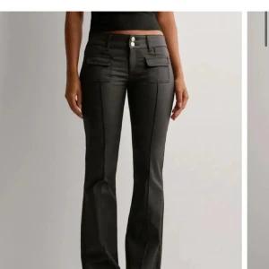 Svarta bootcut byxor med fickor - Säljer ett par svarta byxor med bootcut-snitt och låg midja. Byxorna har dubbla knappar framtill och coola fickor med lock på låren. Snygga sömmar längs benen ger en extra edge. Perfekta för dig som gillar en trendig och stilren look.