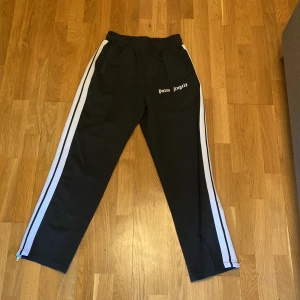 Palm Angels byxor svart - Svarta mjukisbyxor från Palm Angels med vita sidoremsor och logotyp broderad på framsidan. Byxorna har elastisk midja med snörning och dragkedja vid benslut. Snygg streetwear-look och skön passform, perfekta för chill eller häng.