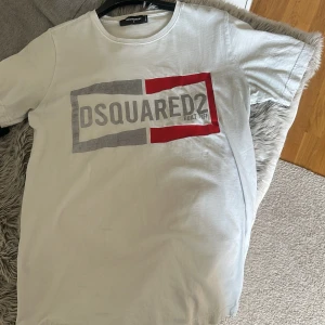 Vit DSQUARED2 t-shirt med tryck - Snygg vit t-shirt från DSQUARED2 i storlek S. Framsidan har ett stort grafiskt tryck med grå och röd ram runt loggan. Loggan är även som en reflex i mörker!  Klassisk rund hals och korta ärmar. Perfekt för dig som gillar streetwear och vill sticka ut med ett coolt märke.