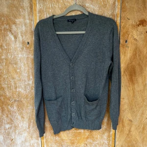 Grå kofta från Urban Research - Mysig grå kofta från Urban Research med v-ringning, knappar framtill och två fickor. Långärmad och ribbade muddar vid ärmslut och nederkant. Perfekt att slänga på sig över en t-shirt eller skjorta för en chill look.