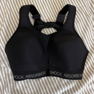 Svart sport-bh från Shock Absorber - Sport-bh i svart från Shock Absorber med breda axelband, justerbar ryggknäppning och meshdetaljer för extra andningsförmåga. Elastiskt band med logga runt om och smidiga spännen både fram och bak. Ej säker på storlek då lapparna klippts bort men lite stor på mig som har xs. Säljs nytt för cirka 500kr.