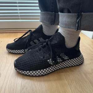 Adidas Deerupt Runner svart/vit - Säljer ett par Adidas Deerupt Runner i svart mesh med vit rutmönstrad sula. Skorna har snörning och en lätt, flexibel ovandel i syntetmaterial. Perfekta för dig som gillar en clean och modern sneakerstil.