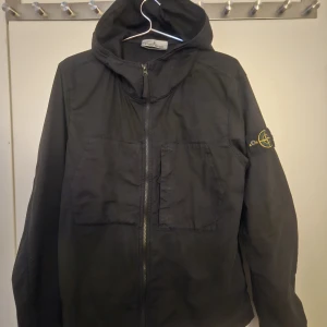 Svart vindjacka från Stone Island - Svart vindjacka från Stone Island med huva och dragkedja framtill. Jackan har två stora bröstfickor och den ikoniska loggan på vänster ärm. Perfekt för dig som gillar streetwear och vill ha en clean look.