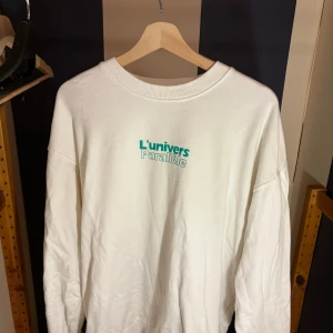 Vit sweatshirt från Vaillent L'univers Parallèle - Vit sweatshirt från Vaillent i storlek L med trycket 'L'univers Parallèle' i turkos och grått framtill. Tröjan har rund halsringning, långa ärmar och en relaxed passform. Perfekt för dig som gillar clean och stilren streetwear.
