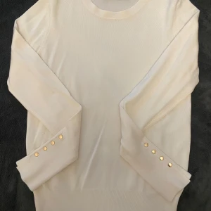 Beige stickad tröja med gulddetaljer - Snygg beige stickad tröja med rund halsringning och långa ärmar. Ärmsluten är dekorerade med fyra guldknappar som ger en extra touch. Perfekt för dig som gillar stilrena och eleganta plagg med en twist.