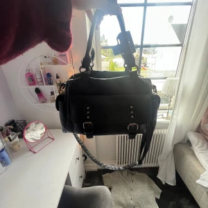 Väska  - Säljer en svart axelväska i skinn med coola detaljer som nitar och spännen framtill. Väskan har både handtag och axelrem, perfekt för att bära på olika sätt. Snygg och rymlig modell som passar till det mesta. Från SHEIN ❤️