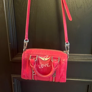 Rosa axelväska Zadig & Voltaire skinn - Cool rosa axelväska i skinn från Zadig & Voltaire med texten 'LOVE' framtill och 'ZADDICTED' på sidan. Väskan har silverfärgade detaljer, dubbla dragkedjor framtill och justerbar axelrem. Perfekt för dig som vill sticka ut med en färgstark accessoar.