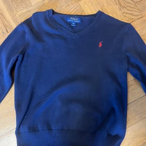 Marinblå stickad tröja Polo Ralph Lauren -  Storlek medium i barn 10- 12 Klassisk marinblå stickad tröja från Polo Ralph Lauren med v-ringning och röd broderad logga på bröstet. Tröjan har ribbade muddar. 