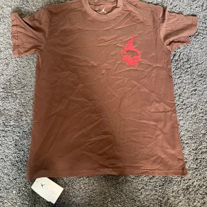 Jordan x Travis Scott brun t-shirt - Brun t-shirt från Jordan x Travis Scott med röd Jumpman-logga och Nike Air-tryck på bröstet. Baksidan har röd text med 'Cactus Jack Highest'. Skön bomullskvalitet och klassisk passform, perfekt för dig som gillar streetwear och snygga collabs.