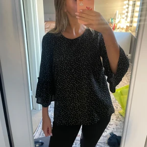 Svart prickig blus med volangärm - Svart blus med små vita prickar och volangdetaljer på de trekvarts-långa ärmarna.