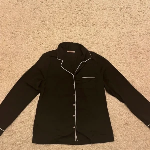 Svart skjorta med vita detaljer Anna Field - Svart långärmad skjorta från Anna Field med klassisk krage och vita kontrastsömmar längs kanterna och vid bröstfickan. Skjortan har knäppning framtill och är tillverkad i mjukt material som känns skönt mot huden. Perfekt för dig som gillar stilren och enkel design.