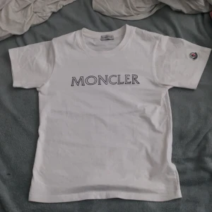 Vit Moncler t-shirt med logga - Vit t-shirt från Moncler med klassisk rund hals och korta ärmar. Stor Moncler-logga tryckt på bröstet och ett broderat Moncler-märke på vänster ärm. Tillverkad i mjuk bomull för skön känsla och clean look. Perfekt för dig som gillar stilrena designer och exklusiva märken.