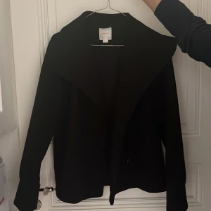Svart kavaj med bred krage - Stilren svart kavaj från Gina tricot med bred krage och enkel knäppning framtill. Jackan har en rak passform och är långärmad, perfekt att slänga på över en t-shirt eller blus. Materialet känns mjukt och har en klassisk look.