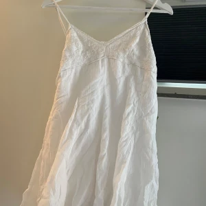 Vit långklänning med spetsdetaljer - Säljer en vit kortklänning från ZARA med tunna axelband och v-ringad spets upptill. Klänningen är ärmlös och har en luftig, flowy passform i mjukt linnematerial. Perfekt för varma dagar och somriga tillfällen. Spetsen ger en romantisk touch och klänningen är superenkel att styla. Helt oanvänd. 