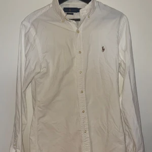 Vit skjorta från Ralph Lauren Slim Fit - Vit långärmad skjorta från Ralph Lauren i slim fit-modell. Klassisk button down-krage, broderad logga på bröstet och knäppning framtill. Skjortan är gjord i bomull och har en stilren och tidlös look.