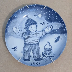 Jultallrik Nr. 21  - 1987 Limited Edition av Paul  Hoff - Unik samlartallrik från 1987, designad av Paul Hoff. Motivet visar en vinterklädd person med fåglar i snölandskap, perfekt för dig som gillar konst och natur. Limited edition från Gustavsberg, Made in Sweden. Snygg att hänga upp i hemmet! Nummer 17 från den välkända serien från Gustavsberg.