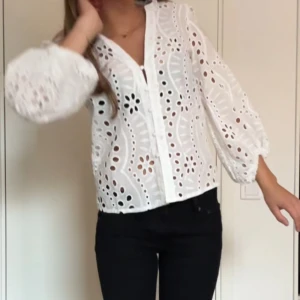 Vit broderad skjorta med v-ringning - Säljer en vit skjorta med v-ringning och broderade detaljer. Skjortan har ett genombrutet mönster med blommiga former och knappar framtill. Ärmarna är långa och puffiga med resår vid ärmslut. Perfekt för dig som gillar romantisk och trendig stil.