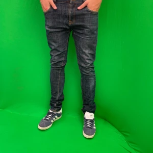 Nudie jeans - Snygga mörkblå jeans från Nudie med klassiska orange sömmar och broderade bakfickor.storlek 31W 34L. Perfekta för dig som gillar en slimmad look. Finns några små hål i fickan.