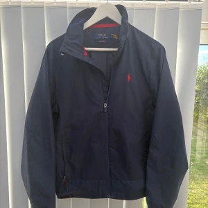 Mörkblå vindjacka Polo Ralph Lauren XS - Snygg mörkblå vindjacka från Polo Ralph Lauren i storlek XS. Jackan har dragkedja framtill, röd logga på bröstet och diskreta vita tryckdetaljer på ärmen. Den är perfekt för blåsiga dagar. Klassisk krage och enkel design med röda inslag. Skicket är som nytt 