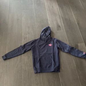 Mörkblå Adidas Originals hoodie XS - Säljer en mörkblå hoodie från Adidas Originals i bomull med coolt färgglatt dinosaurie-tryck på ryggen och liten rosa detalj framtill. Tröjan har huva med snörning, känguruficka och långa ärmar. Perfekt för dig som gillar streetstyle och unika prints.