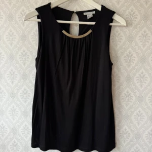 Svart ärmlös topp med gulddetalj - Svart ärmlös topp från H&M med snygg guldfärgad metall-detalj vid halsringningen och en liten öppning i ryggen. Toppen har en rak passform och är tillverkad i mjukt, stretchigt material som känns skönt mot huden. Rökfritt hem.