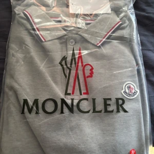 Grå Moncler pikétröja XXL - Snygg grå pikétröja från Moncler med stor logga framtill och broderad patch på bröstet. Kragen har röda och vita ränder som ger en sportig vibe. Perfekt för dig som vill ha en stilren och trendig look.