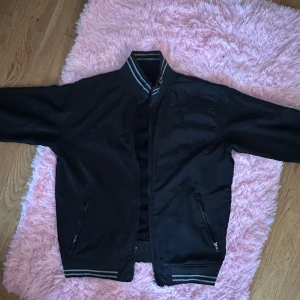 Svart bomberjacka med randiga muddar - Svart bomberjacka med klassiska randiga muddar vid krage, ärmslut och nederkant. Jackan har dragkedja framtill och två fickor med dragkedja. Snygg och enkel modell som passar till det mesta. Perfekt för dig som gillar en stilren look.