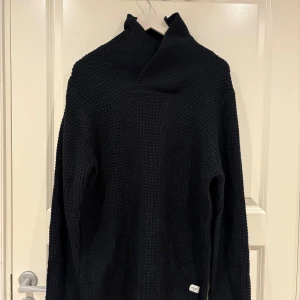 Svart stickad polotröja - Säljer en svart stickad polotröja med grov struktur och hög, bred krage. Tröjan har lång ärm och en avslappnad passform, perfekt för kyliga dagar. Enkel att matcha med jeans för en stilren look.