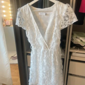 Vit blommig kortklänning från Vila - Superfin vit kortklänning från Vila med blommigt spetsmönster och v-ringad omlottfront. Klänningen har korta ärmar i transparent spets🤍🤍🤍🤍