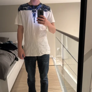 Vit t-shirt från Marcelo Burlon  - Snygg vit t-shirt med korta ärmar och ett fett blått fågeltryck över axlarna och halsen. Rund halsringning och normal passform. Materialet känns som mjuk bomull, perfekt för chill dagar. Trycket ger en unik vibe och sticker ut på ett stilrent sätt. Lite stor som du kan se, jag är 185cm och väger 75kg