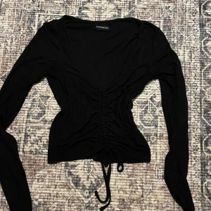 Svart ribbad topp med snörning Even&Odd - Svart långärmad topp från Even&Odd med ribbad struktur och snygg v-ringning. Toppen har snörning framtill som ger en cool rynkad effekt och croppad känsla. Perfekt att styla med jeans eller kjol för en trendig look.