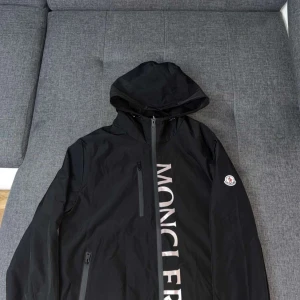 Svart vindjacka från Moncler med huva - Svart vindjacka från Moncler med stor logga längs dragkedjan och klassisk patch på ärmen. Jackan har huva, vattentäta dragkedjor och en clean look. Perfekt för dig som gillar stilren streetwear med premiumkänsla.