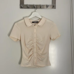 Beige blus med rynk från Bik Bok - Säljer en beige blus från Bik Bok i storlek XS. Toppen har korta ärmar, klassisk krage och knappar hela vägen framtill. Snygg rynkdetalj längs knappraden ger en cool och trendig look. Perfekt till jeans eller kjol för en fräsch stil. (Aldrig använd)