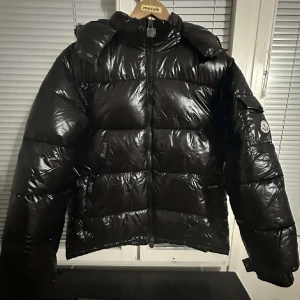 Svart Moncler jacka - Säljer en svart, glansig pufferjacka från Moncler med huva och dragkedja framtill. Jackan har en klassisk Moncler-logga på ärmen och stora, vadderade sektioner som ger en riktigt fet look. Perfekt för kalla dagar när du vill hålla stilen.