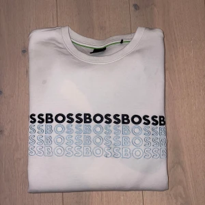 Vit sweatshirt från BOSS med tryck - Vit sweatshirt från BOSS med svart och ljusblått BOSS-tryck över bröstet. Klassisk rund halsringning och långa ärmar. Skön passform och tillverkad i mjuk bomull, perfekt för en clean och stilren look.