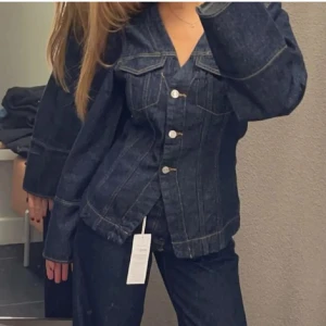 Mörkblå jeansjacka  - Snygg mörkblå jeansjacka med markerade sömmar och silverfärgade knappar framtill. Jackan har klassisk krage, två bröstfickor och lång ärm. Perfekt för dig som gillar en tidlös och cool look.