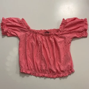 Rosa offshoulder topp från Gina Tricot - Supersöt rosa offshoulder topp från Gina Tricot med broderade hålmönster och volangdetaljer. Croppad modell med resår i både ärmslut och nederkant för en snygg passform. Perfekt för dig som vill ha en feminin och trendig look. Går även att ha den on-shoulder.