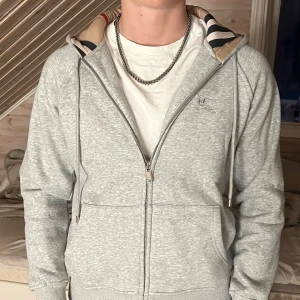 Grå Burberry hoodie med dragkedja - Säljer en grå hoodie från Burberry i storlek M. Tröjan har dragkedja, snörning vid huvan och Burberrys klassiska rutiga mönster på insidan av huvan. Mjuk bomullskänsla och ribbade muddar vid ärmslut och nederkant. Perfekt för dig som gillar stilrena och exklusiva plagg.