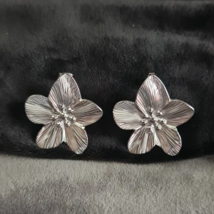 Silvriga blomformade örhängen - Stora örhängen i form av blommor med fem kronblad, tillverkade i blankt silvrigt material. De har detaljerade ådringar och små kulor i mitten som ger en cool och unik look. Perfekta för dig som vill sticka ut med ett statement-smycke.