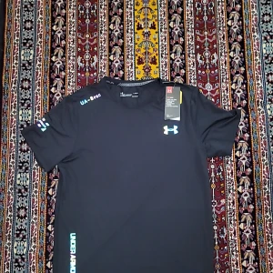 Svart Under Armour HeatGear t-shirt - Svart t-shirt från Under Armour med tryckta loggor i vitt och blått på bröstet, ärmen och nedtill. Modellen är kortärmad och har en rund hals. Tillverkad i lätt och stretchig polyester med HeatGear-teknologi som håller dig sval under träning. Perfekt för dig som gillar sportig stil.
