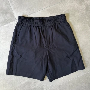 Svarta shorts från Arket, storlek 46 - Svarta shorts från Arket med elastisk midja och raka ben. De har sidfickor fram och två diskreta bakfickor. Yttertyget är en mix av 50% polyester och 50% ull, fickfodret är 100% bomull. Perfekta för chill dagar och enkel stil. B348