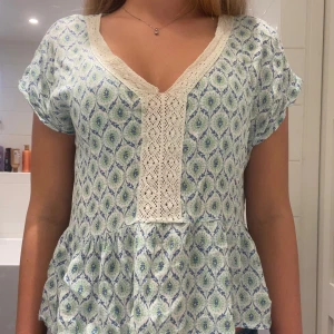 Blommig blus med spetsdetaljer - Supersöt blus i ljusblått och grönt med v-ringning och breda spetsdetaljer framtill. Blusen har korta ärmar, volang nertill och ett luftigt, flowy material som känns perfekt för varma dagar.
