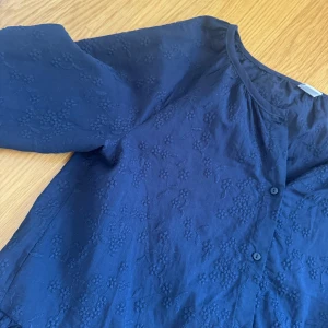 Mörkblå blus med broderade blommor - Supersöt mörkblå blus med broderade blommor och volang nertill. Blusen har v-ringning, knappar framtill och långa ärmar med lätt puff. Perfekt för dig som gillar romantiska detaljer och en avslappnad men ändå stilsäker look.