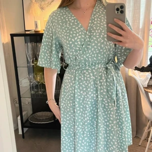 Mintgrön omlottklänning med mönster - Superfin mintgrön omlottklänning med vita prickar, knytband i midjan och v-ringad hals. Klänningen har korta, vida ärmar och ett flowigt fall. Perfekt för dig som gillar ljusa färger och ett lekfullt mönster.